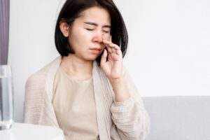 A woman is sitting on a couch with her hand on her nose, possibly experiencing sinus discomfort or seeking relief with CBD.