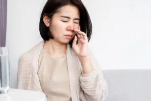 A woman is sitting on a couch with her hand on her nose, possibly experiencing sinus discomfort or seeking relief with CBD.