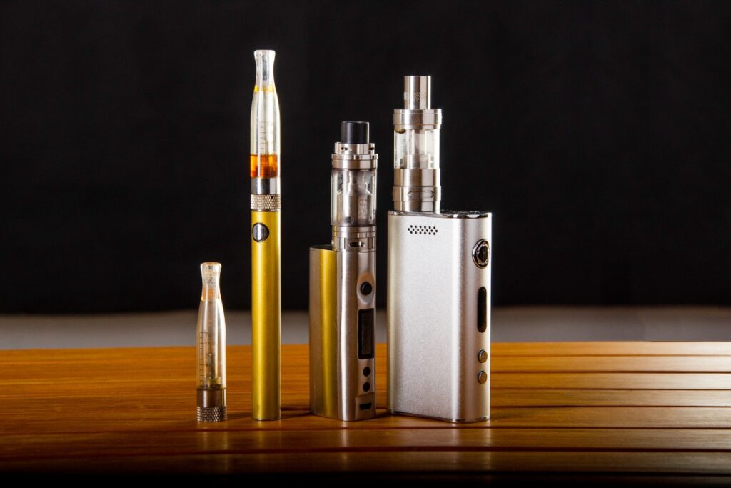 On a wooden table, there is a collection of e-cigs and vaporizers, including an assortment of vape starter kits.