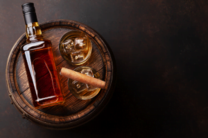 Bottle of whiskey and a glass with ice next to a cigar on a wooden barrel.