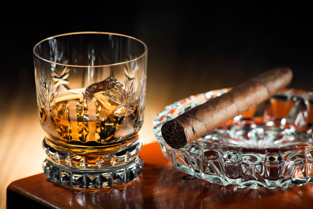 A glass of whiskey with ice next to a cigar on an ashtray.