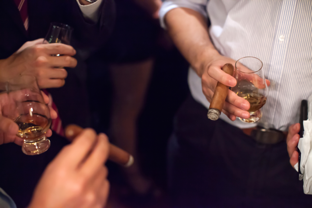 People socializing while holding glasses of spirits and a cigar, discussing cigar pairing tips.