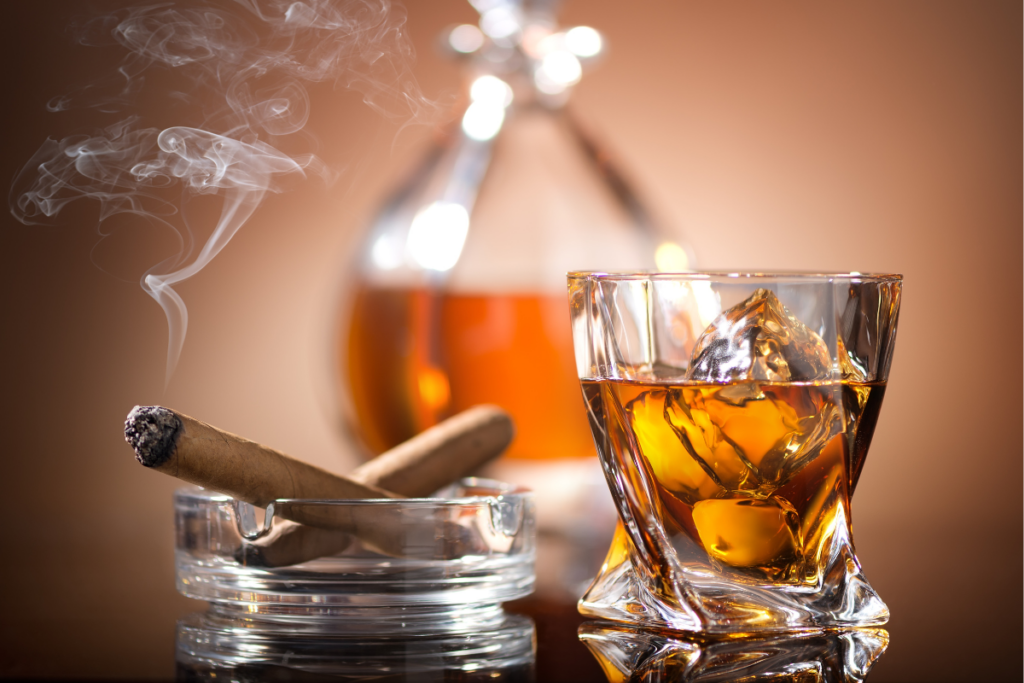 Cigar tips: A glass of whiskey with ice, an ashtray with a smoking cigar, and a bottle in the background, perfectly illustrating the art of cigar pairing.