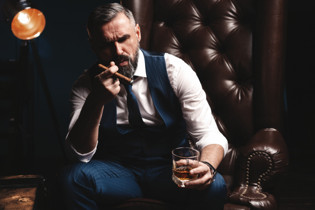 A man in a vest seated on a leather chair, holding cigar and a glass of whiskey.
