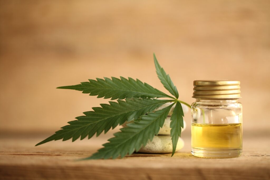 An organic cannabis leaf rests beside a small glass jar of CBD oil on a wooden surface, set against a soft, beige background.
