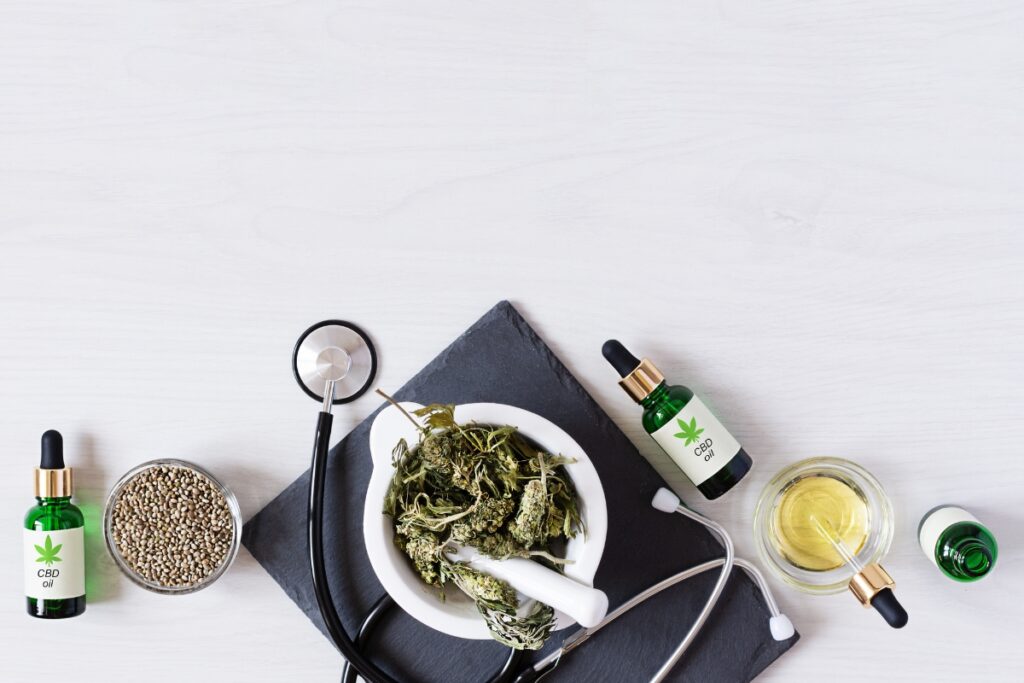 Top view of a workspace with organic cbd oil bottles, hemp leaves in a bowl, seeds, and a stethoscope on a white wooden surface.