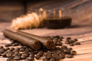 Two cigars lie side by side on a wooden surface, surrounded by scattered coffee beans. A glass container filled with a dark liquid is blurred in the background, setting the perfect scene for an exquisite cigar and coffee pairing.