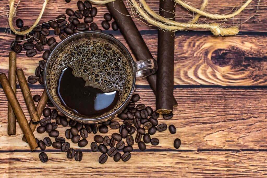 A cup of black coffee on a wooden surface, surrounded by coffee beans, cinnamon sticks, rolled up dark paper, and an invitation to a sophisticated cigar and coffee pairing experience.