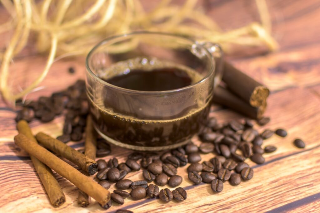 A clear glass cup of black coffee, ideal for a cigar and coffee pairing, is surrounded by coffee beans and cinnamon sticks on a wooden surface.