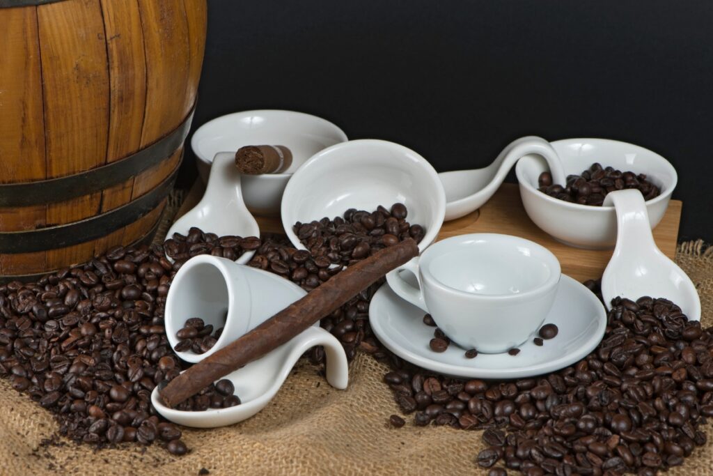 White ceramic coffee cups, saucers, and spoons with scattered coffee beans on a burlap surface; a wooden barrel is partially visible on the left, inviting thoughts of a perfect cigar and coffee pairing.