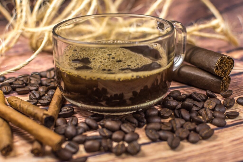 A clear glass cup filled with black coffee is surrounded by coffee beans, two cigars, and cinnamon sticks on a wooden surface, exemplifying the perfect cigar and coffee pairing.