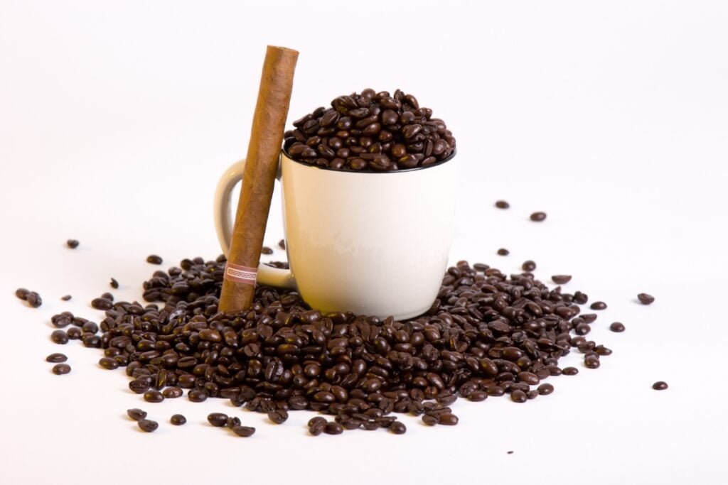 A white mug filled with coffee beans, surrounded by more coffee beans, and a cigar leaning against the mug on a white background—a perfect cigar and coffee pairing.