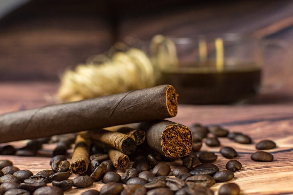 Three cigars rest on a surface adorned with scattered coffee beans, illustrating the perfect cigar and coffee pairing; a glass cup filled with a dark beverage completes the scene in the background.