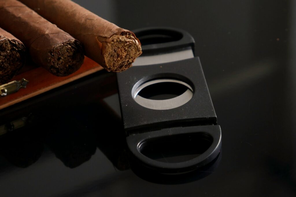 Three cigars, part of tobacco history, resting on a wooden holder next to a black cigar cutter on a reflective surface.