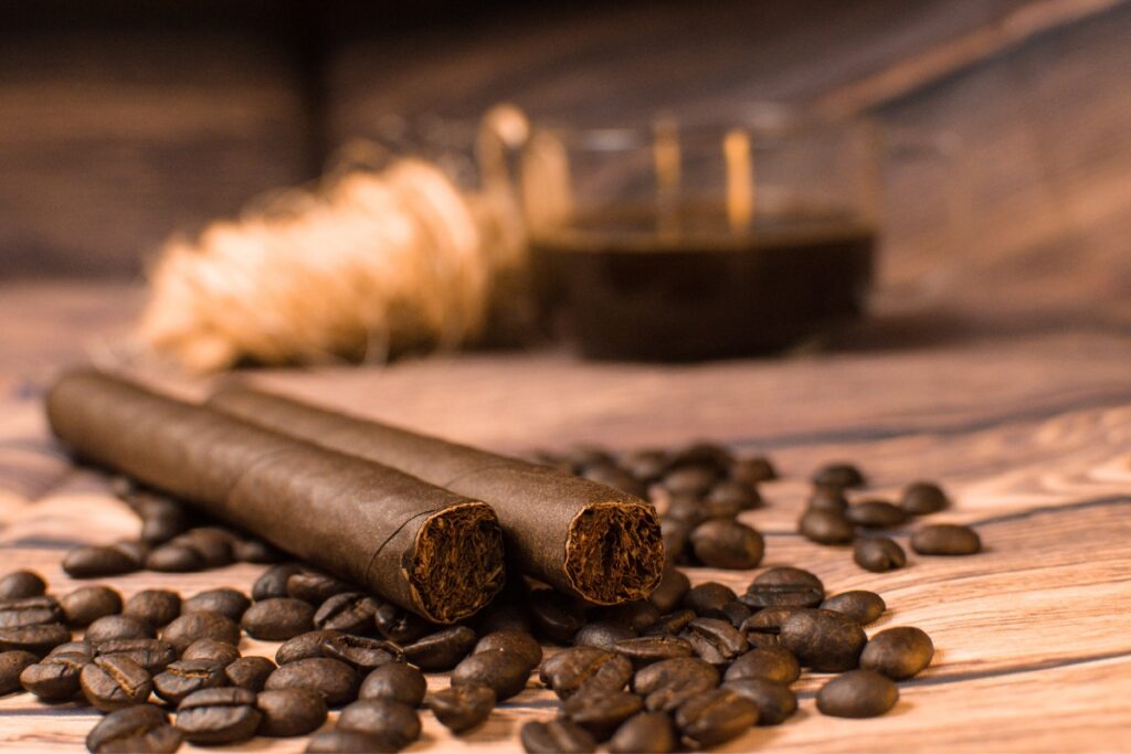 Two hand-rolled cigars, steeped in tobacco history, resting on a bed of coffee beans, with a glass of whiskey and wheat sheaf in the background on a wooden surface.