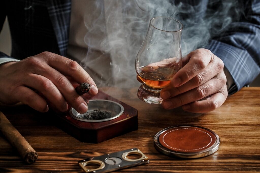 A person holding a cigar and a glass of whiskey, with smoke swirling above a wooden table filled with tobacco history-related accessories.