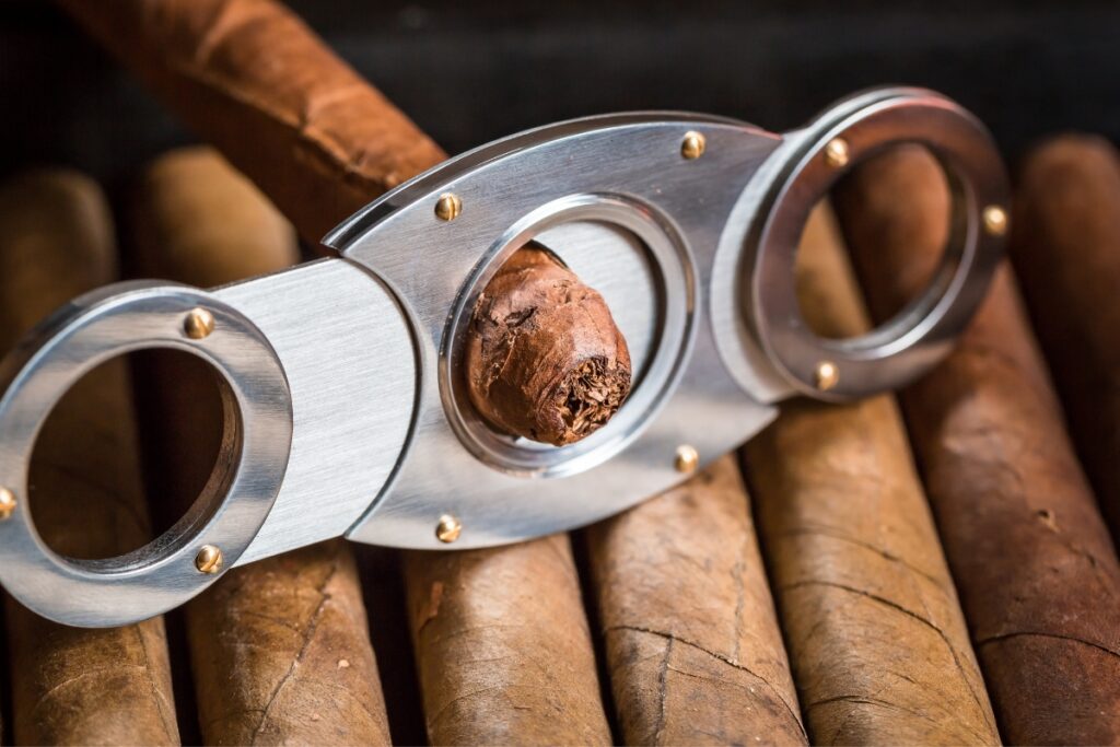 Close-up of a shiny metal cigar cutter positioned over a cigar in a box filled with multiple cigars, illustrating an aspect of tobacco history.