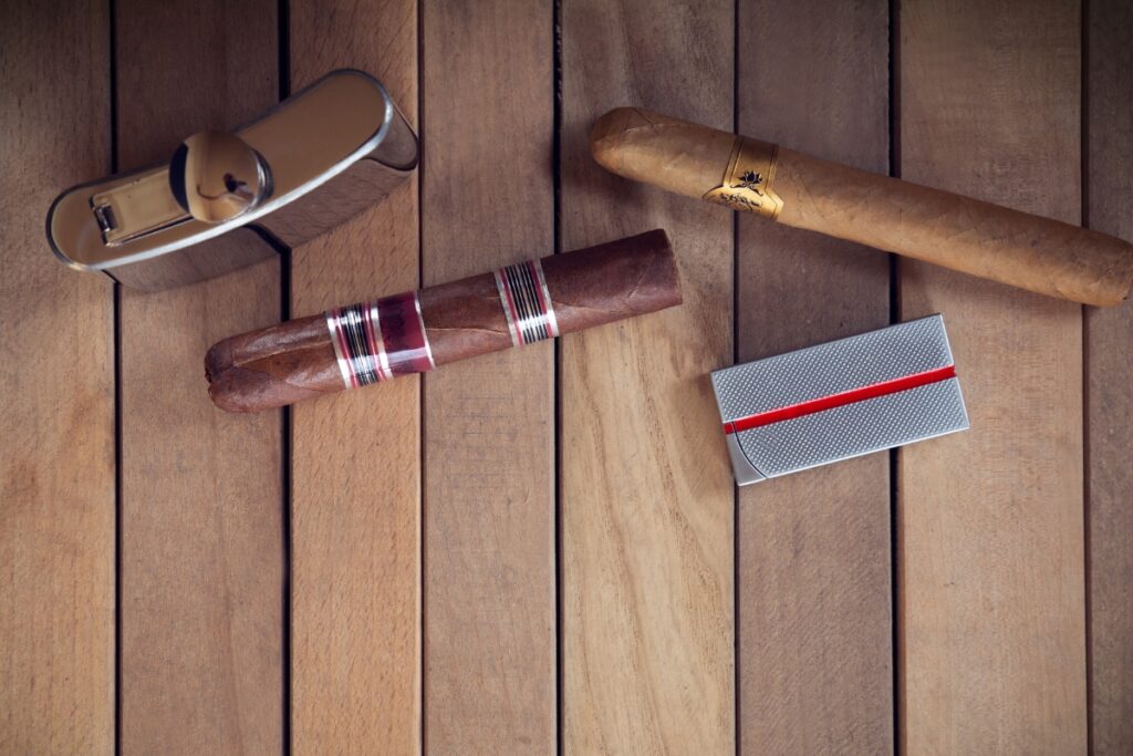 Two cigars, a cigar cutter, and a lighter arranged on a wooden surface, showcasing elements of tobacco history.