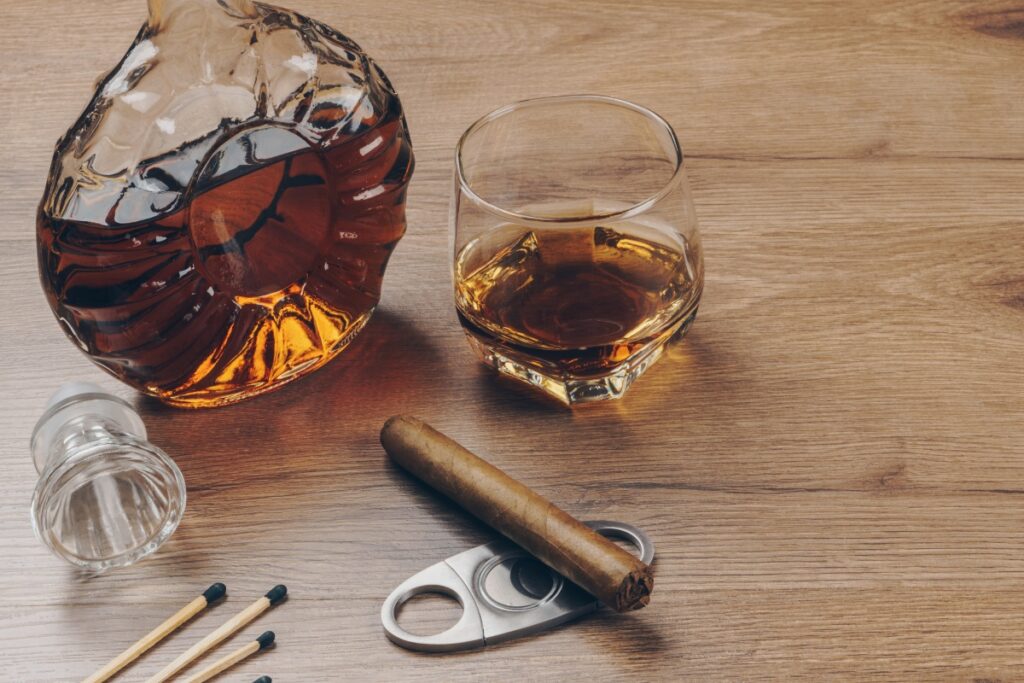 A bottle of whiskey, a glass half-filled with whiskey, a cigar representing tobacco history, matches, and a cigar cutter on a wooden surface.