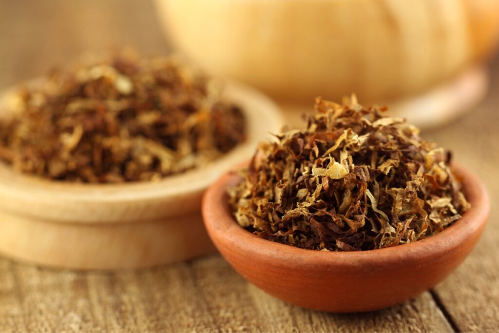 Dried tobacco leaves in two small wooden bowls on a wooden table, adhering to tobacco industry regulations, with a blurred background featuring a whole tobacco leaf.