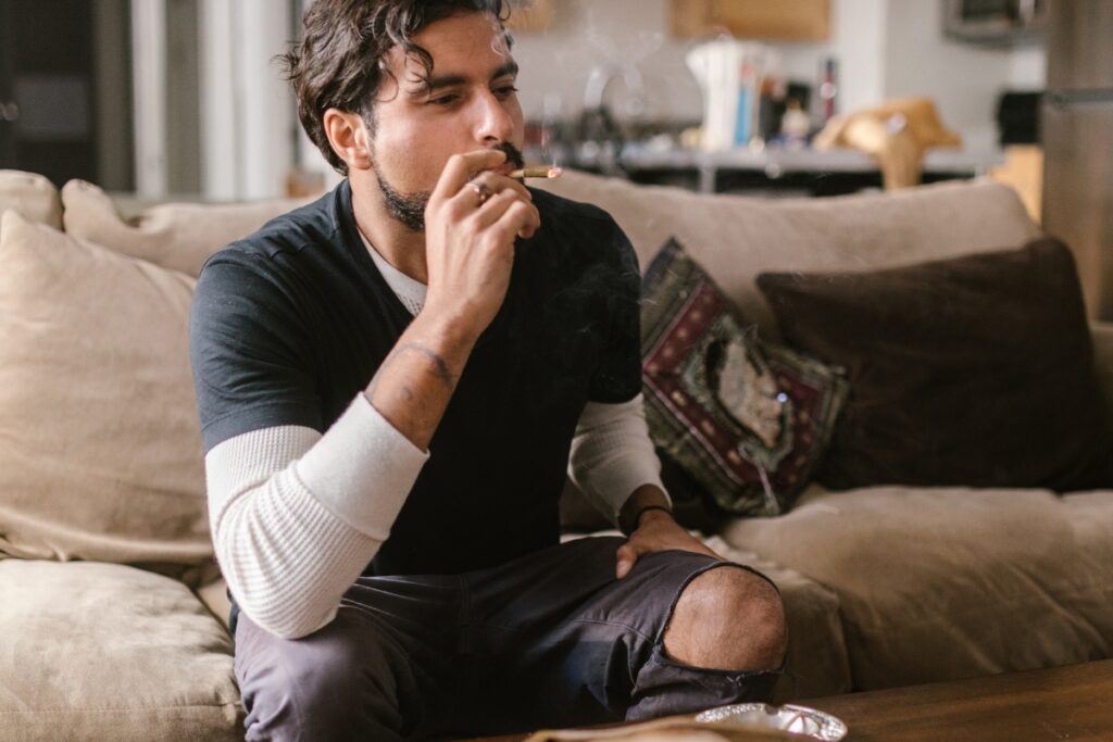 A person with a beard is sitting on a beige couch in their home smoking lounge, enjoying a cigarette. They are wearing a black shirt over a white long-sleeve shirt, with some smoke visible around their head.