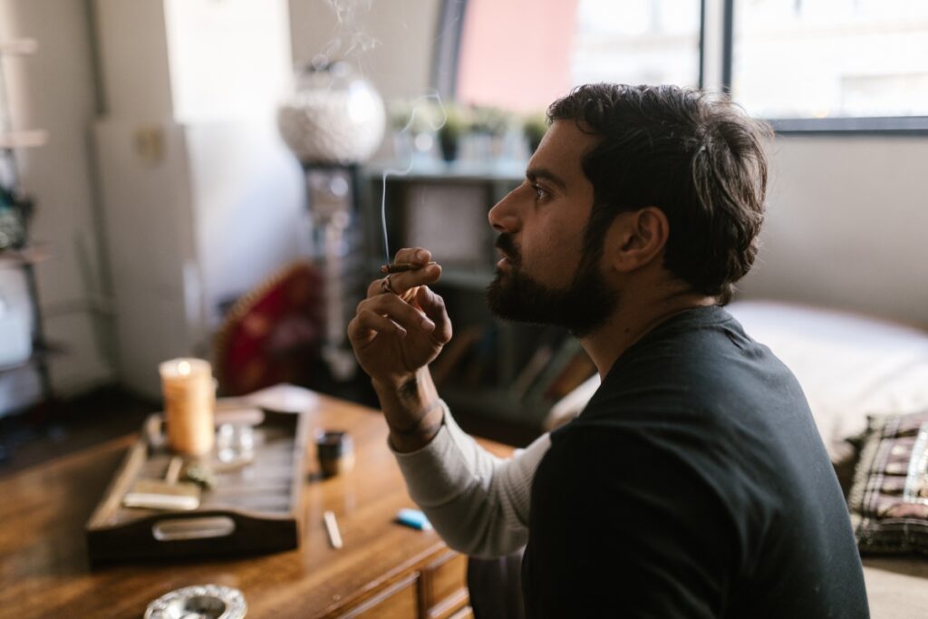 A man with a beard sits indoors in what appears to be a home smoking lounge, holding a lit cigar close to his mouth. He appears to be in deep thought. Various items are scattered on the wooden table in front of him.