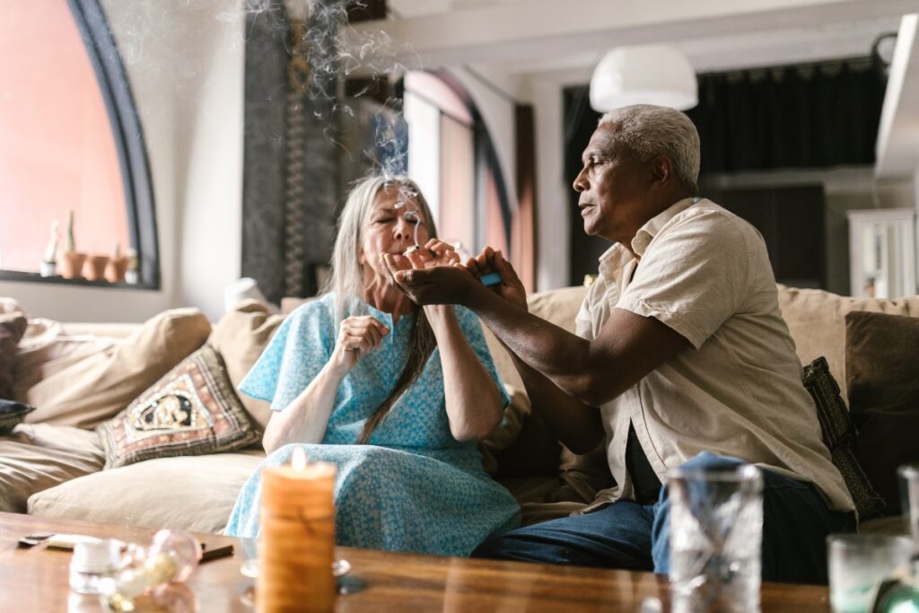 An elderly woman is being assisted by an elderly man in lighting a cigarette while sitting on a couch in what feels like a cozy home smoking lounge. The room has a smoky ambiance, with the soft glow of a lit candle on the table.