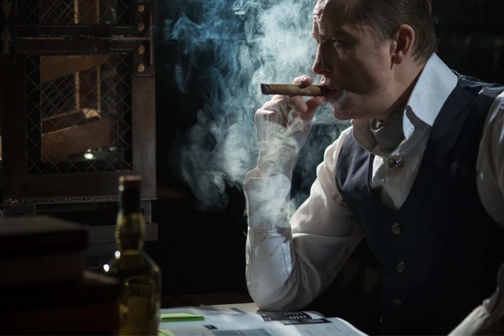 A person in a formal vest and white shirt enjoys a cigar in their home smoking lounge near a table with books and a bottle, smoke swirling around.