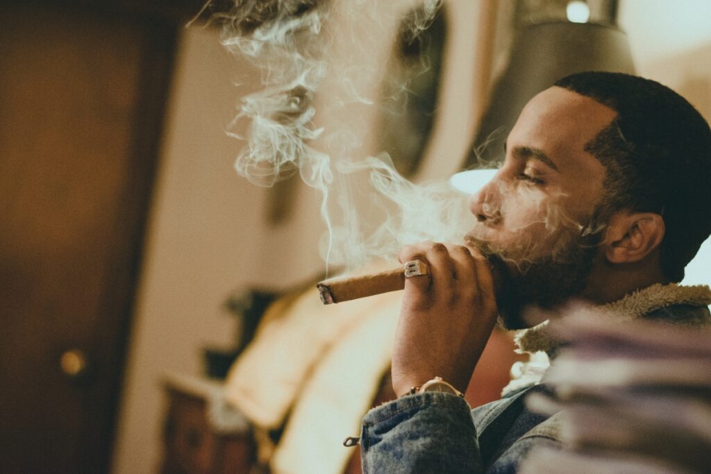 A man is sitting in his home smoking lounge, a cigar in hand. Smoke swirls around his face as he exhales.