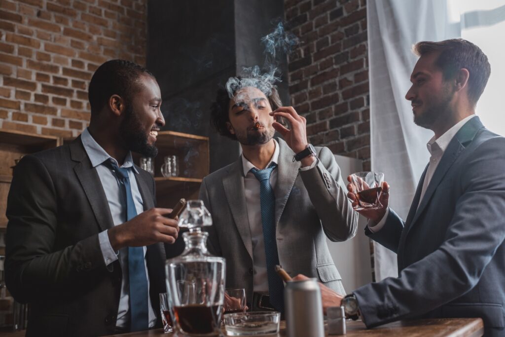 Three men in suits are enjoying drinks and cigars in a stylish home smoking lounge with exposed brick walls. One man is smoking a cigar, while the others hold glasses of whiskey. A decanter is on the table.