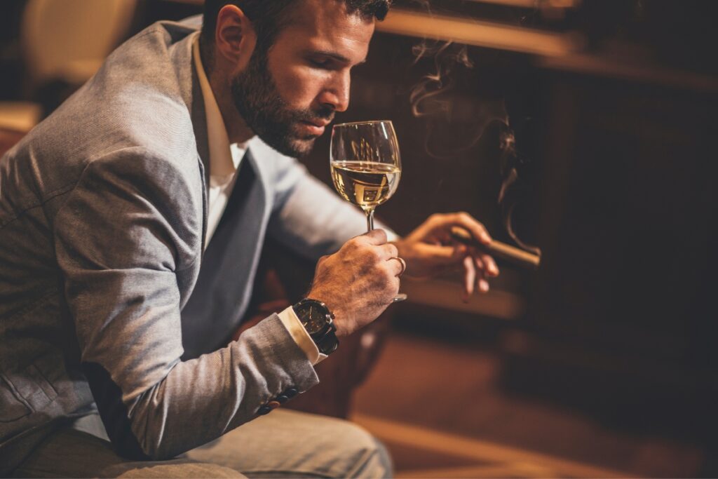 A man in a light grey blazer is sitting in his home smoking lounge, holding a wine glass in one hand and a cigar in the other.