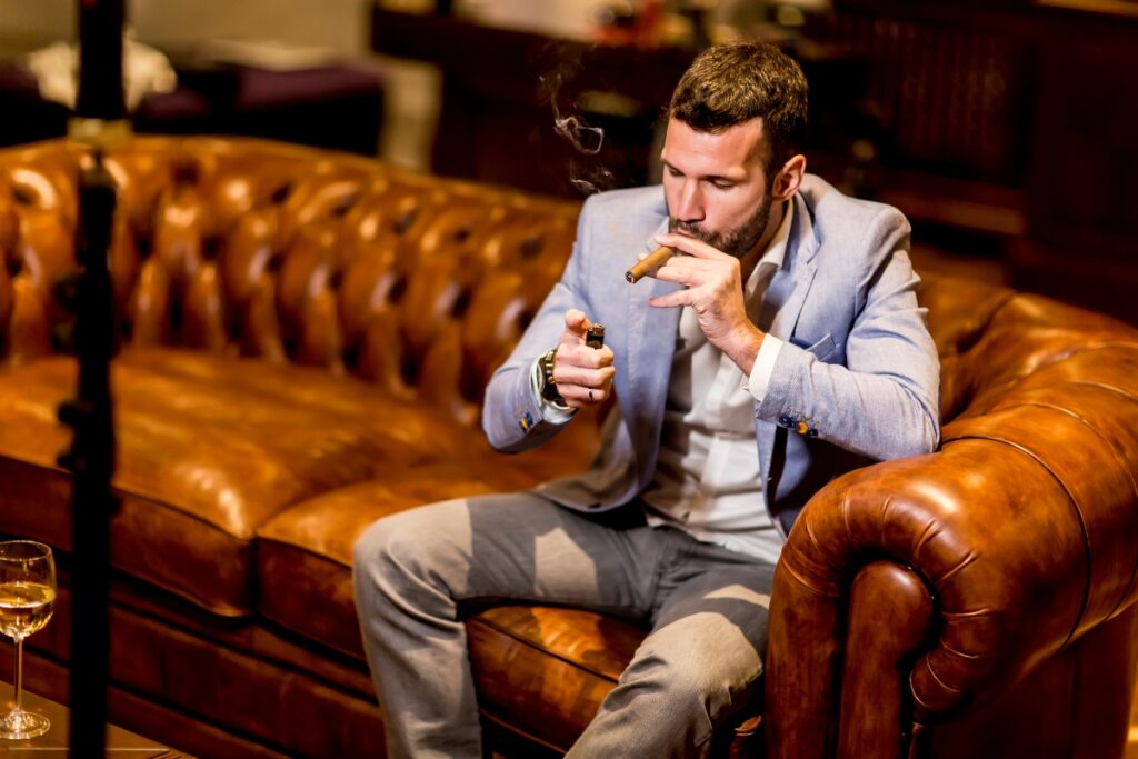 A man sits on a brown leather sofa in his home smoking lounge, lighting a cigar with a small torch lighter, dressed in a blue blazer and gray pants. A glass of drink is on the table beside him.