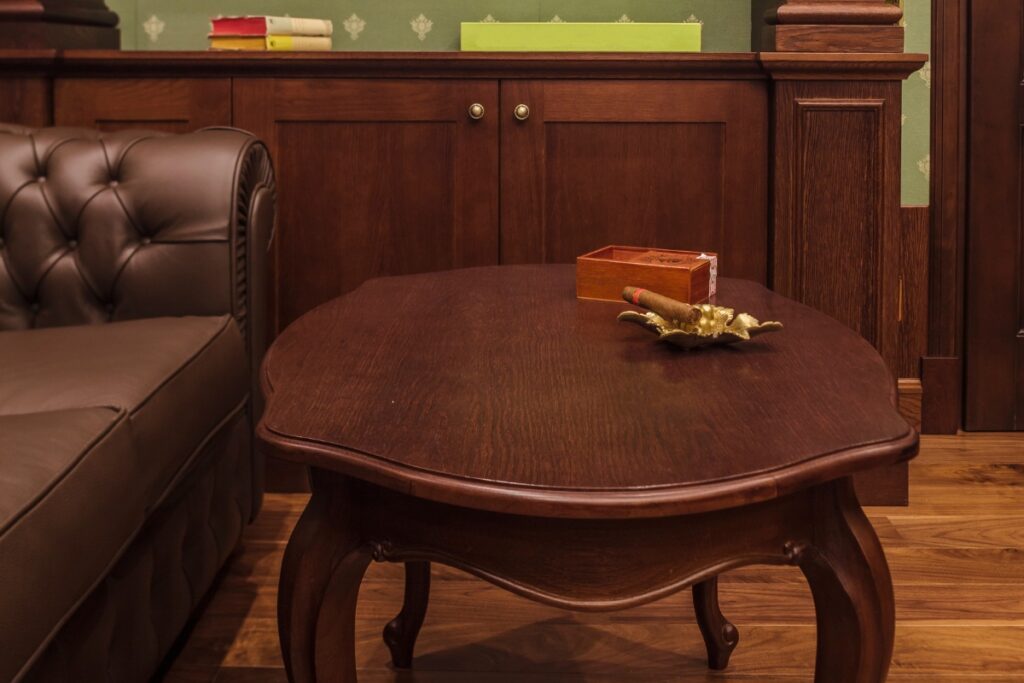 A wooden coffee table with an ornate edge sits next to a tufted leather sofa in the cozy home smoking lounge. The table holds a small wooden box and a decorative item resembling a golden leaf. There are shelves and cabinets behind.