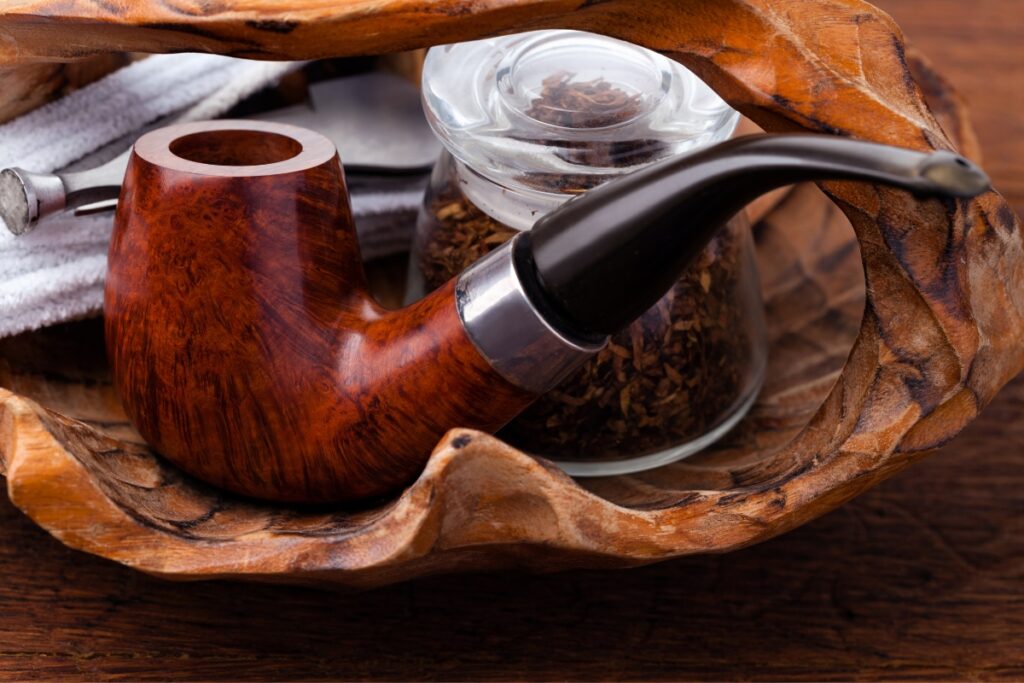 A wooden smoking pipe and a glass jar filled with tobacco are placed in a carved wooden bowl, with a white cloth partially visible—a nod to modern pipe smokers seeking classic elegance.