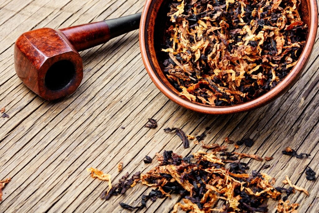 A wooden tobacco pipe and a bowl filled with tobacco sit on a rustic wooden surface, some loose tobacco scattered around, evoking a sense of nostalgia for modern pipe smokers.