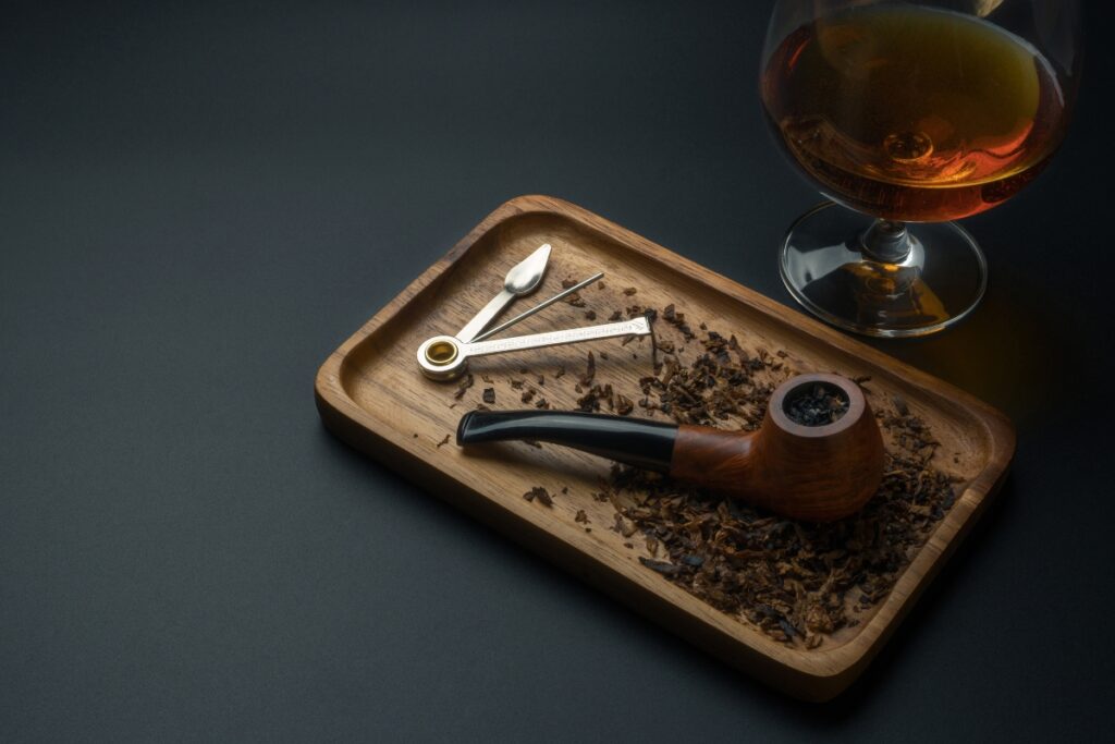 A wooden tray holds a tobacco pipe, pipe tool, and loose tobacco. For modern pipe smokers appreciating the classics, a glass of amber-colored liquid stands beside the tray.