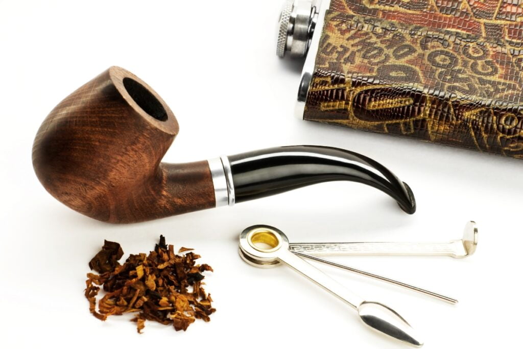 A wooden tobacco pipe, a small pile of tobacco, a pipe tool, and a patterned leather-covered flask are placed on a white surface, capturing the aesthetic cherished by modern pipe smokers.