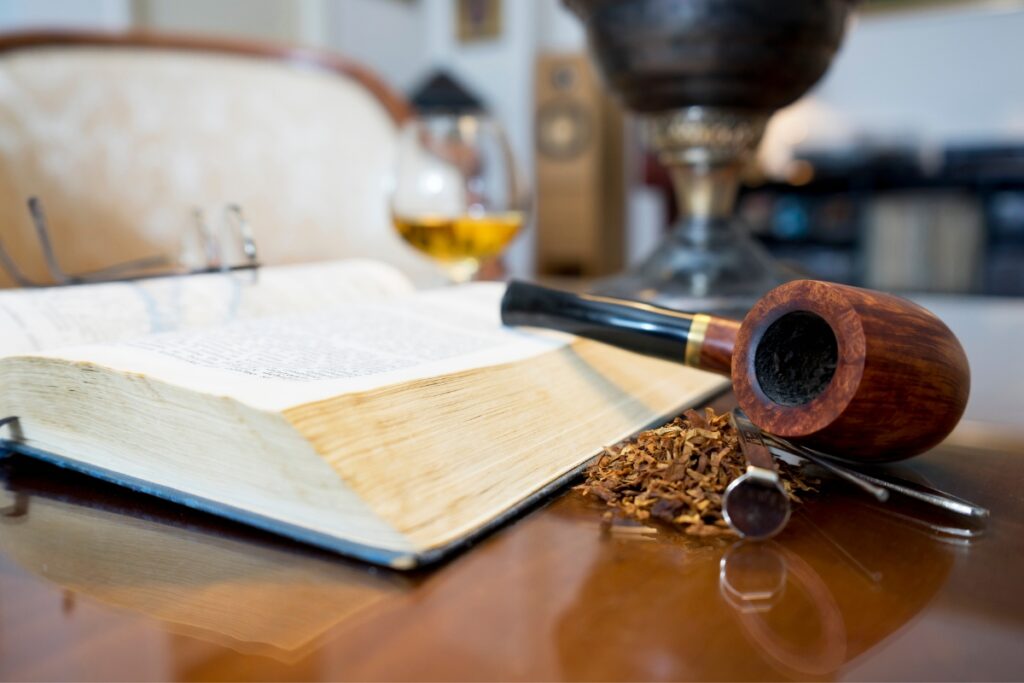 Open book with a pair of eyeglasses on top, a tobacco pipe, and loose tobacco placed on a wooden table, perfect for modern pipe smokers.