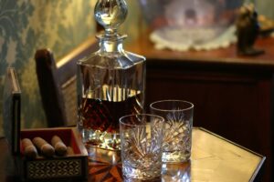 A crystal decanter filled with a dark liquid stands on a wooden table next to two empty crystal glasses and an open box containing four cigars, perfectly setting the scene for indulging in fine craft spirits.