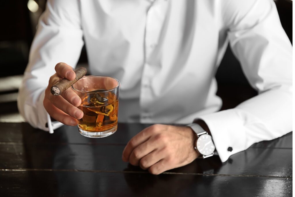 A person in a white shirt holds a glass of craft spirits with ice in one hand and a lit cigar in the other, resting on a dark wooden table—ideal for those who appreciate fine cigar pairings.