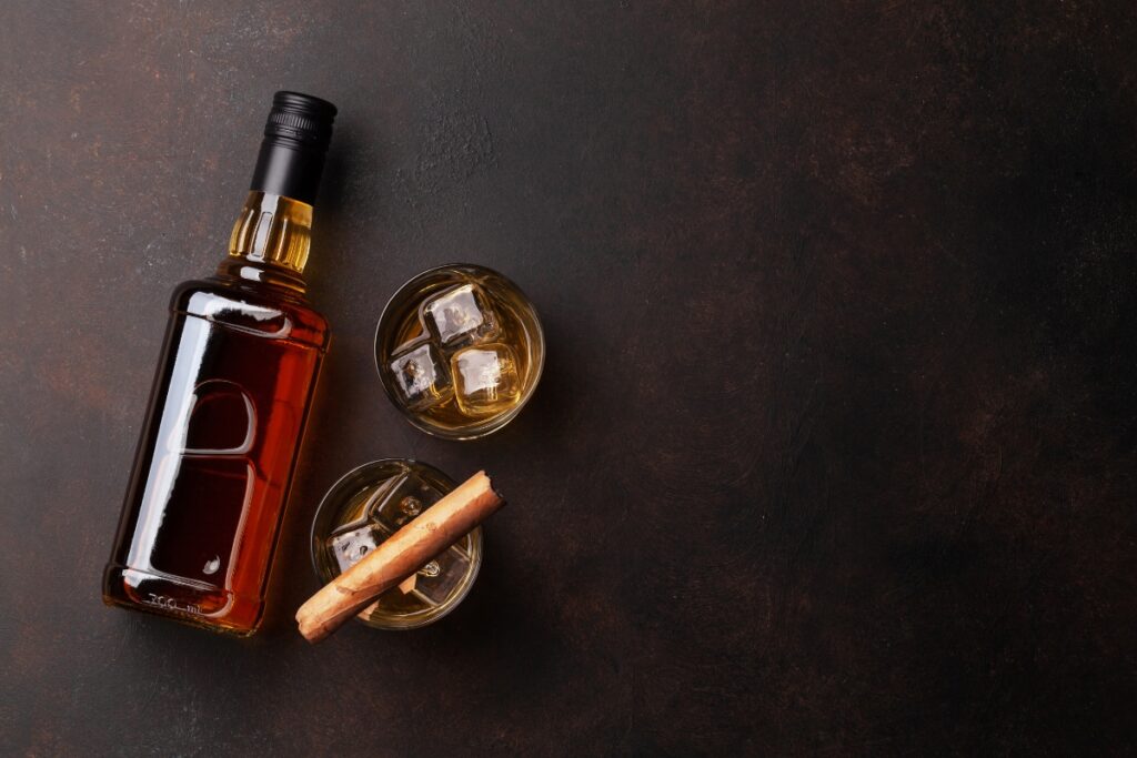 A bottle of craft spirits lies on a dark surface next to two glasses of whiskey with ice, one glass also containing cinnamon sticks—an ideal setup for refined cigar pairings.