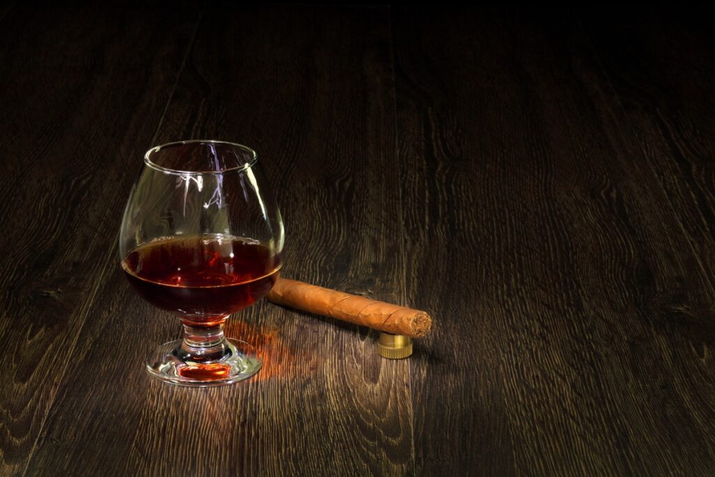 A glass of dark craft spirits and a cigar rest on a dark wooden surface, offering an exquisite Cigar Pairing.