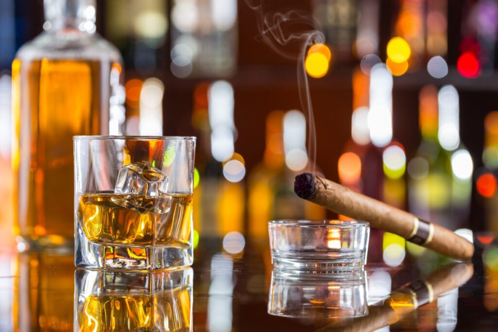 A glass of whiskey with ice, a burning cigar in an ashtray, and a bottle of craft spirits are placed on a reflective surface with a blurred background of colorful lights.