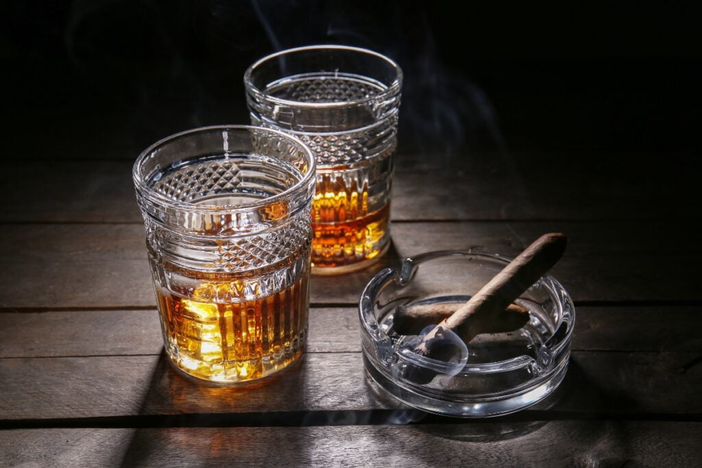 Two glasses of craft spirits with ice sit on a wooden table alongside a cigar resting in an ashtray, all under dim lighting—a perfect setting for refined cigar pairings.