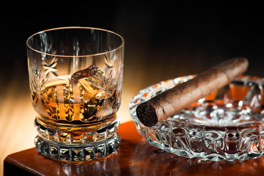 A glass of craft spirits with ice sits next to a lit cigar on a crystal ashtray on a wooden surface, creating the perfect setting for cigar pairings.