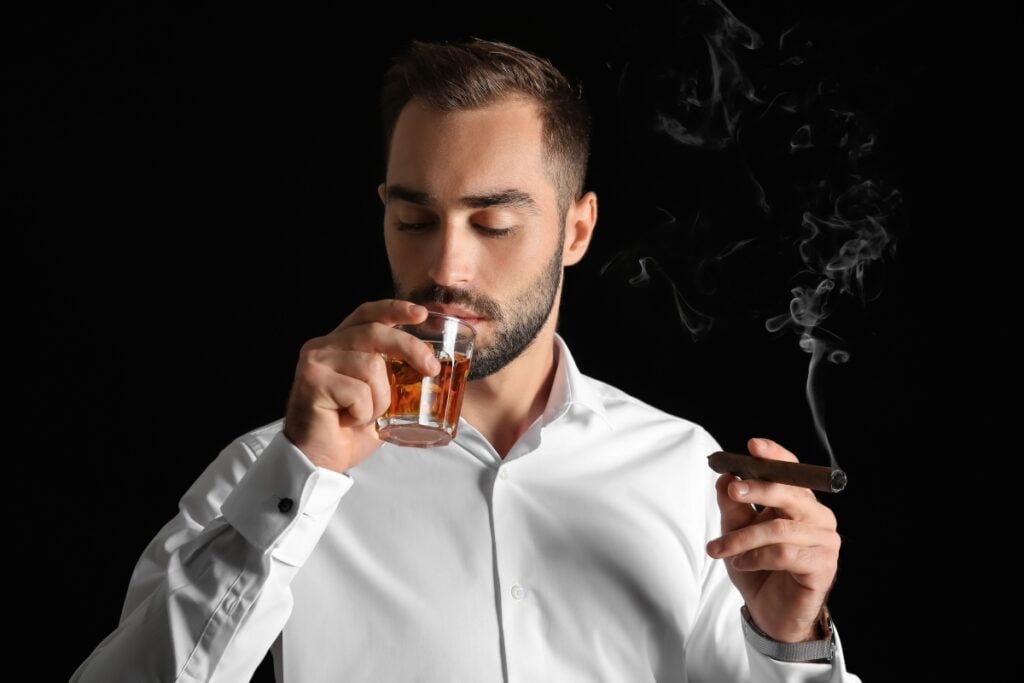 A man in a white shirt sips a craft spirit from a glass while holding a lit cigar, with smoke rising from the cigar against a black background, showcasing perfect cigar pairings.