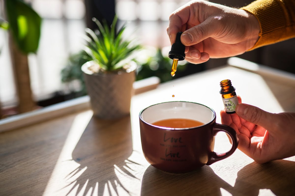 A person drops liquid from a CBD wellness dropper bottle into a mug of tea on a wooden table with a potted plant in the background.