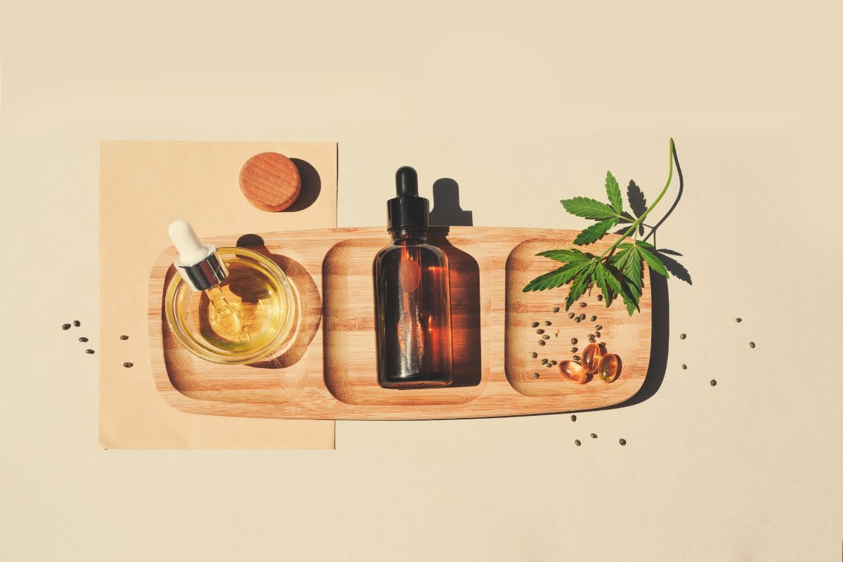 A wooden tray holds a glass dropper bottle of golden CBD wellness liquid, an amber bottle with a dropper, a green leaf, seeds, and gel capsules against a light background.