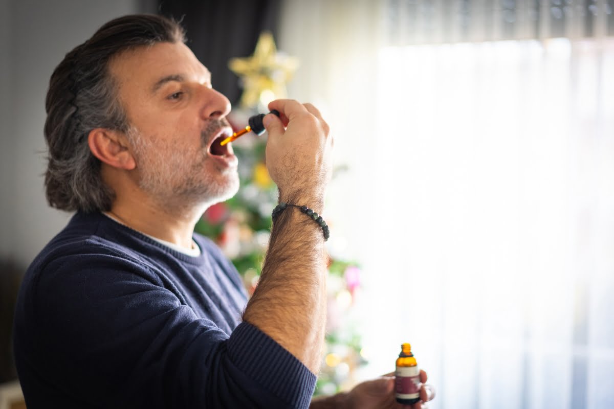 A man with gray hair uses a dropper to place CBD wellness liquid into his mouth, standing near a decorated Christmas tree.