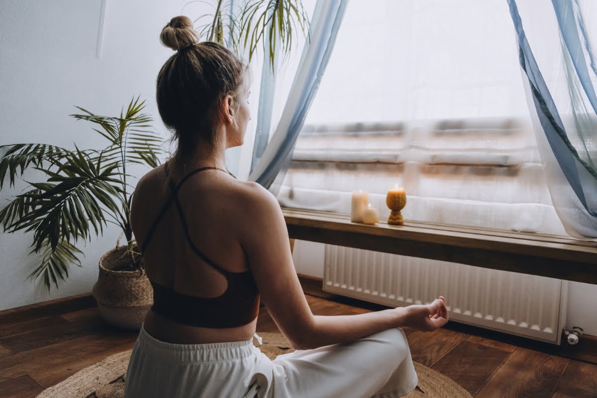 A woman in a cross-back sports top and loose pants meditates on a mat in front of a window with lit candles, surrounded by indoor plants, embracing the tranquility of her CBD wellness routine.